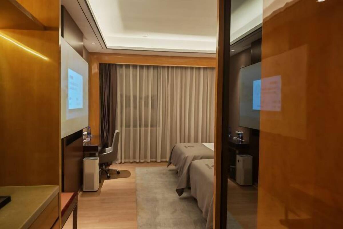 A hotel room with a desk, two beds, a TV, and a large window with curtains.