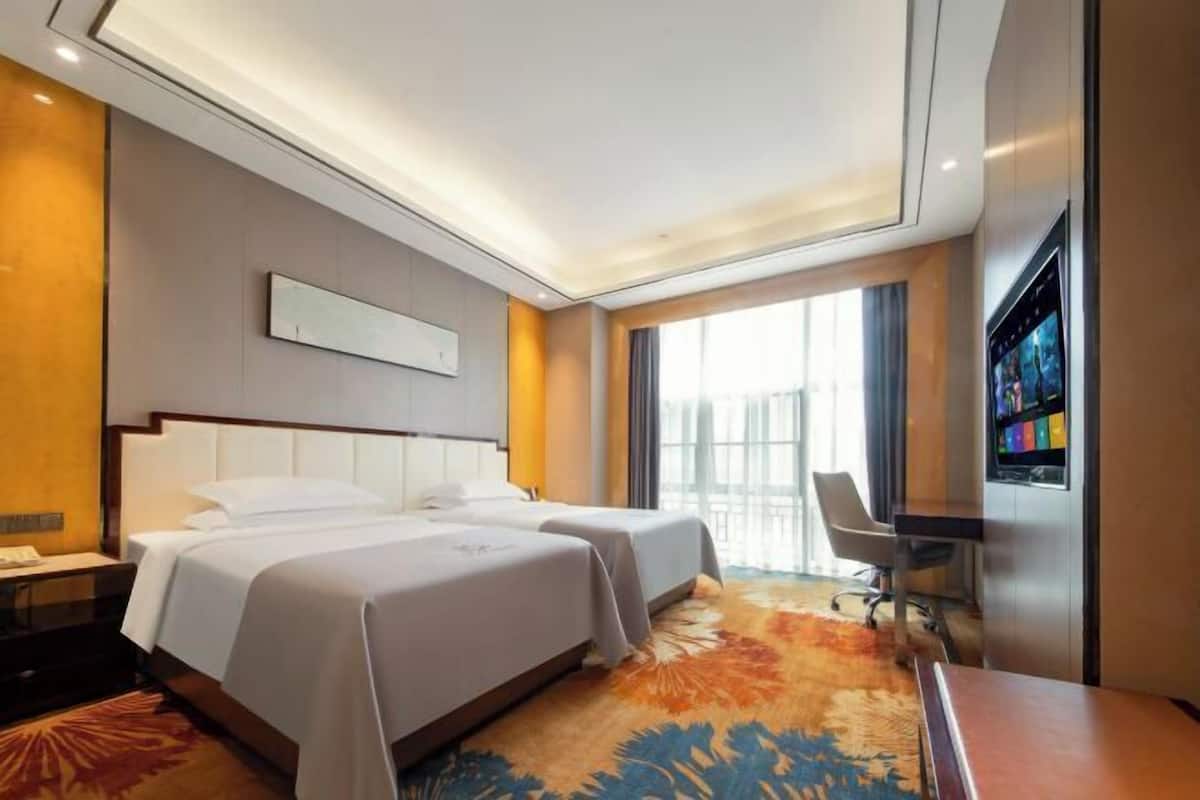 A hotel room with two beds, a desk, a chair, a TV, and a large window.