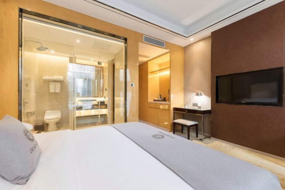 A hotel room with a bed, a desk, a TV, and a bathroom visible through a glass door.