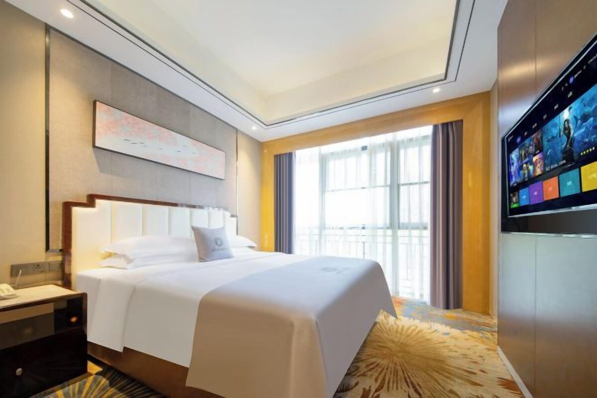 A hotel room with a large bed, a flat-screen TV, a desk with a lamp, and a window with curtains.