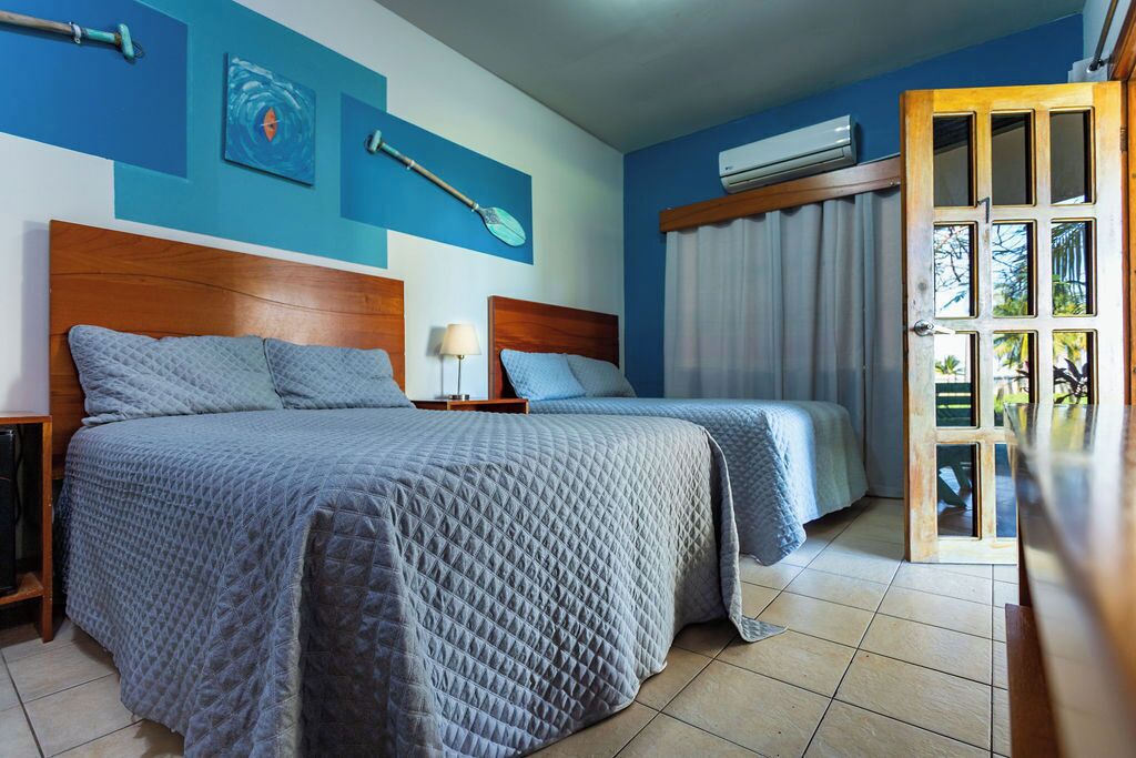 Habitación de hotel con dos camas, un cabecero de madera, una mesita de noche, una lámpara, una ventana con cortinas y un equipo de aire acondicionado.