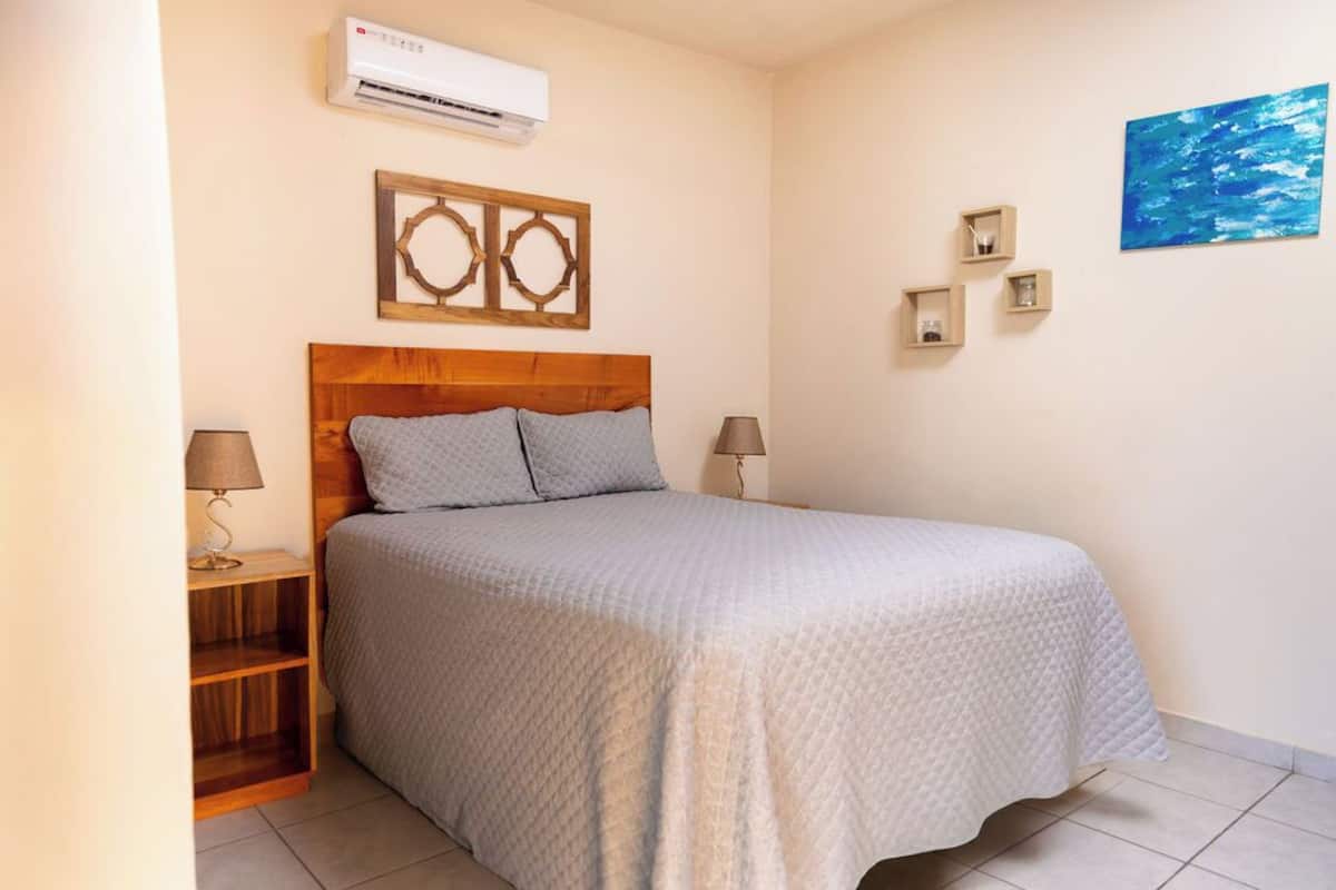 A bedroom with a wooden headboard, a bed with a quilted bedspread, two bedside lamps, a wooden nightstand, a wall-mounted air conditioner, and a blue abstract painting.