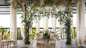 Decoración para una boda al aire libre con terraza de madera, cortinas blancas y arreglos florales.