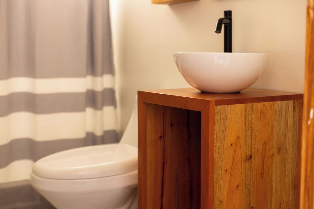 Ein Badezimmer mit Toilette, einem Holzkästen und einem weißen Waschbecken mit schwarzem Wasserhahn.