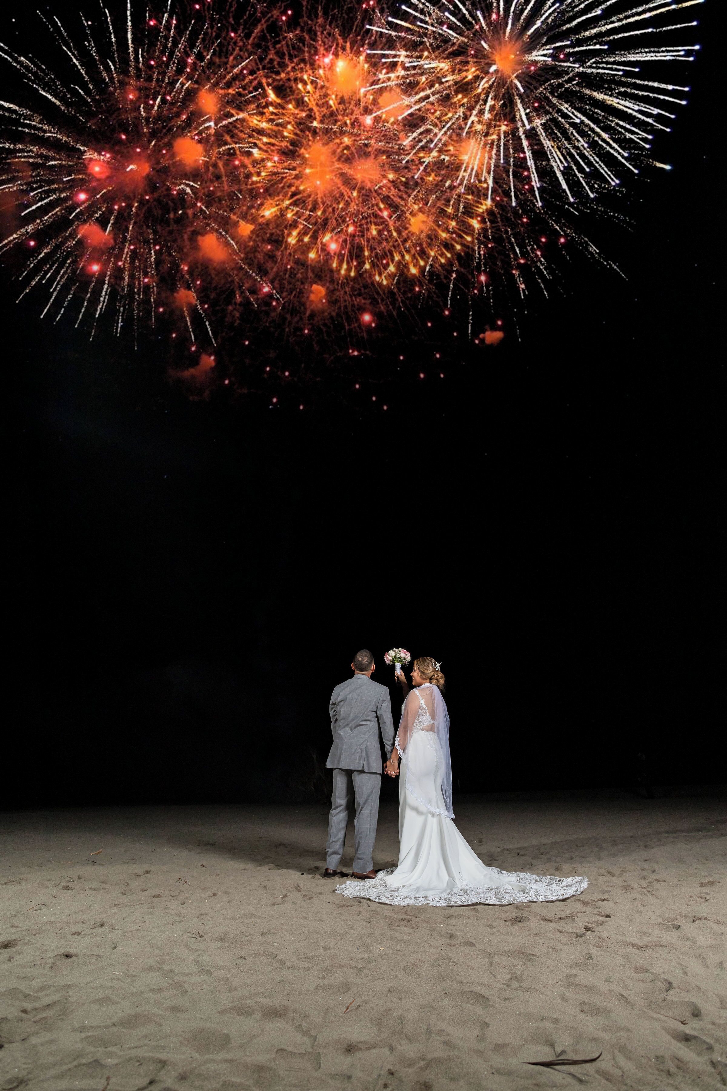 A couple standing on a sandy beach with fireworks in the background.