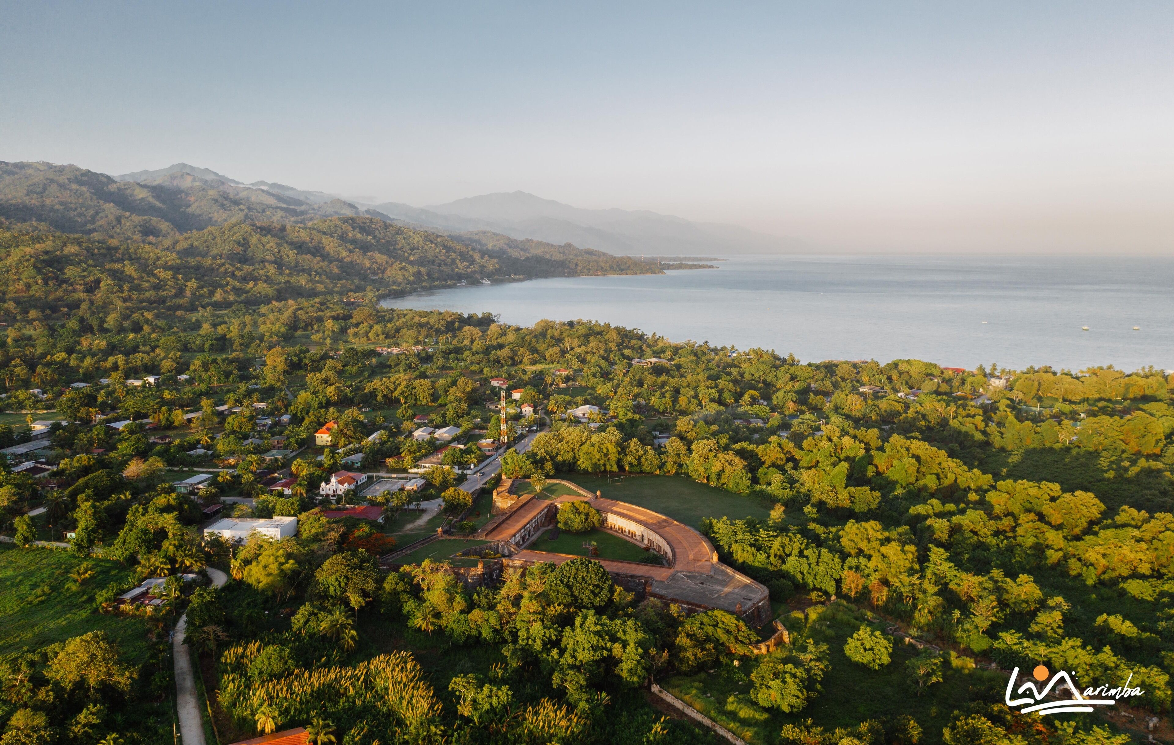 An aerial view of a coastal area with a mix of residential buildings, greenery, and a large body of water.