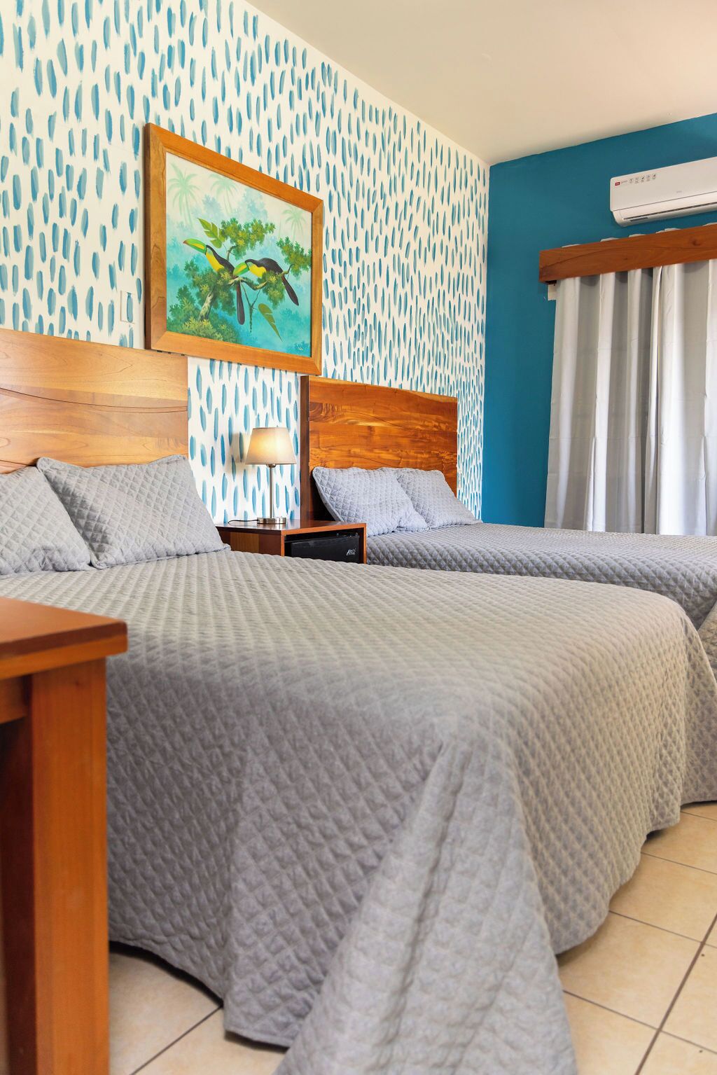 Habitación de hotel con dos camas, cabecera de madera, mesita de noche con lámpara, un cuadro de una escena tropical y una ventana con cortinas.