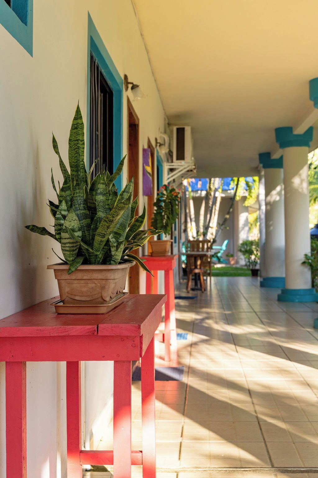 Área externa coberta com uma planta em vaso sobre um banco vermelho, chão de azulejos e uma fileira de cadeiras e mesas ao fundo.