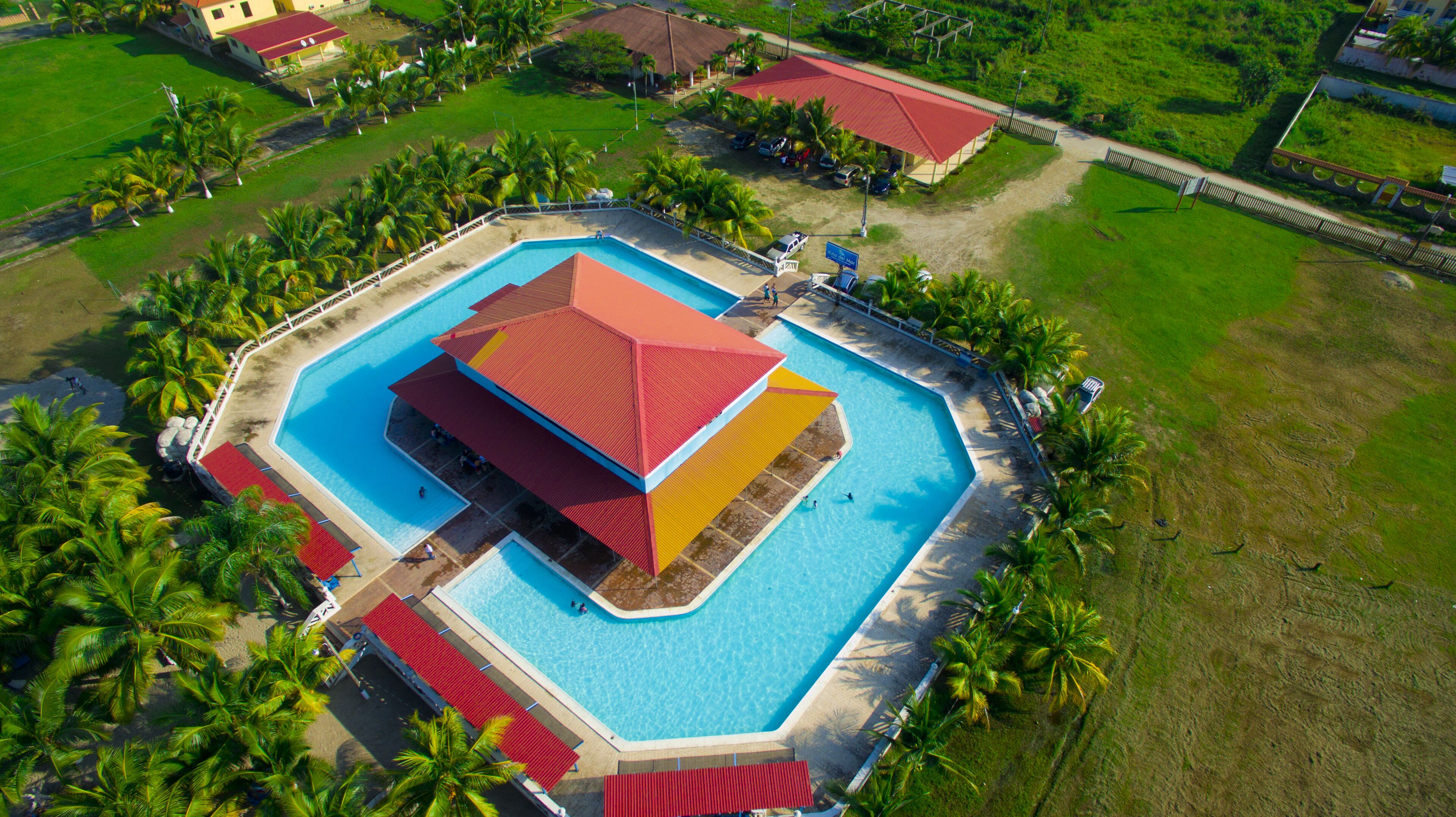 Vista aérea de un resort con una piscina grande, palmeras y un edificio de techo rojo.