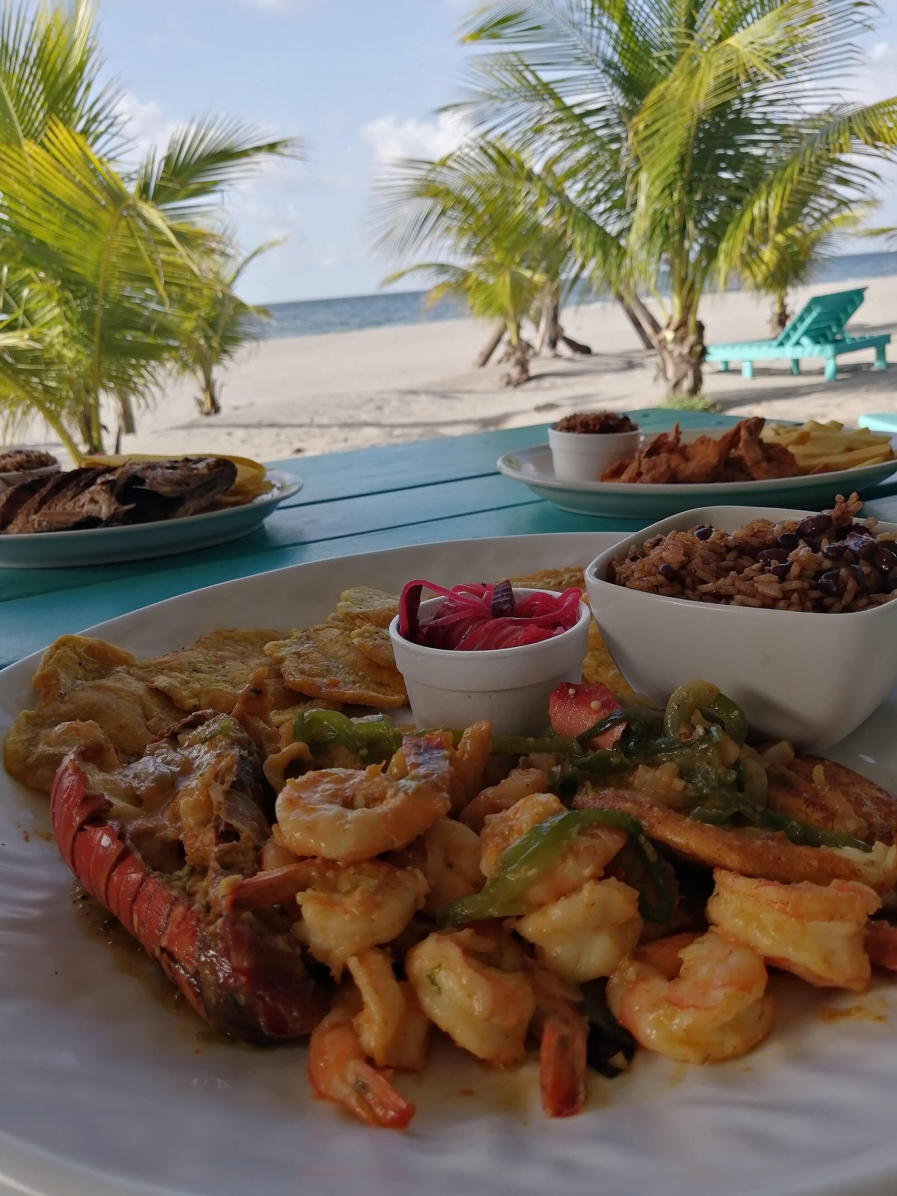 A beachside meal with shrimp, vegetables, and sides on a table with a view of the beach and palm trees.