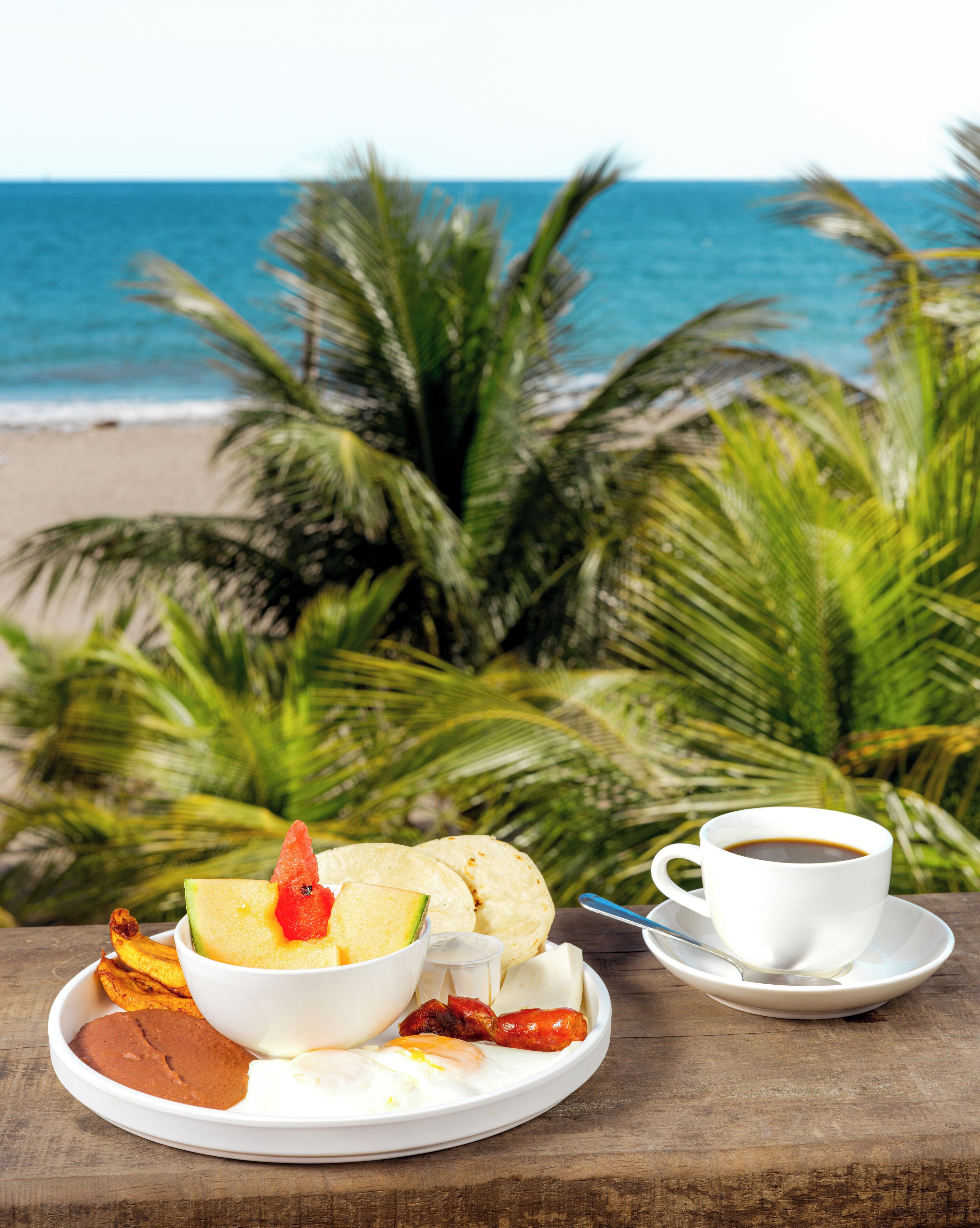 A meal with a cup of coffee on a wooden table with a view of the ocean and palm trees in the background.