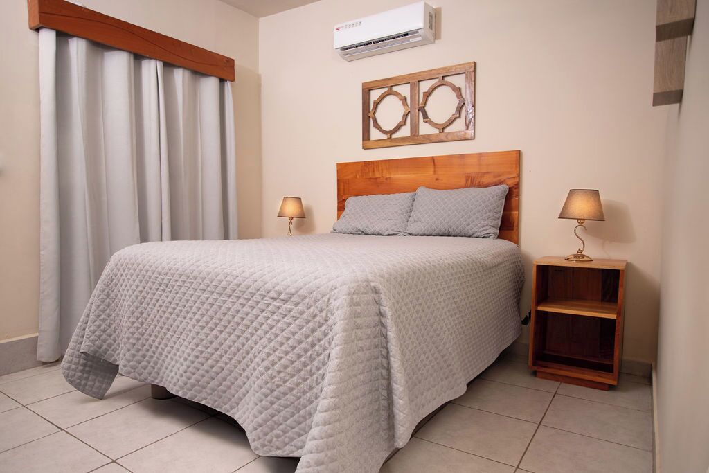 Una cama bien hecha con cubrecama acolchado, dos lámparas de noche, un cabecero de madera, un aire acondicionado instalado en la pared y un adorno de madera sobre la cama.