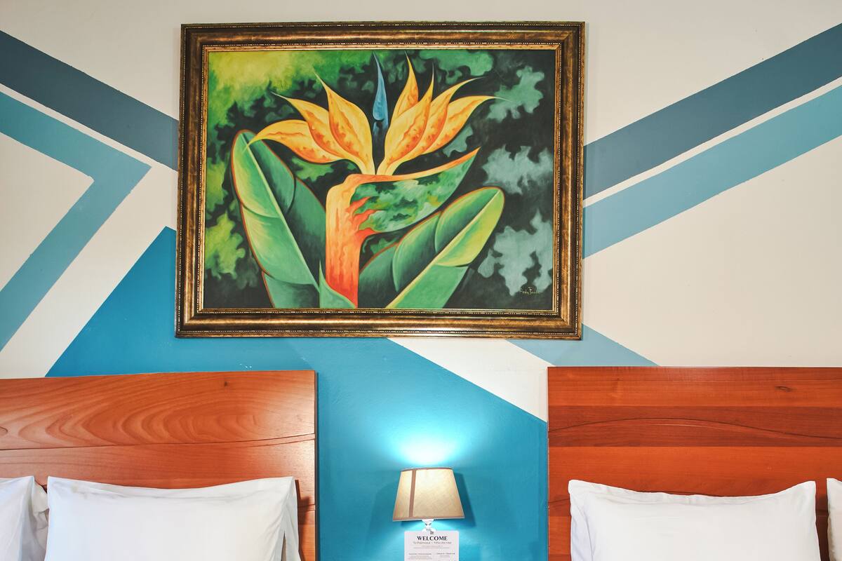 A hotel room with two beds, a nightstand with a lamp, a welcome sign, and a painting on the wall.