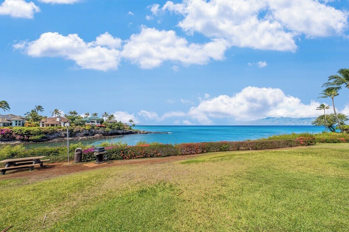 A grassy area with a view of a beach and ocean, houses in the background, and a picnic table.