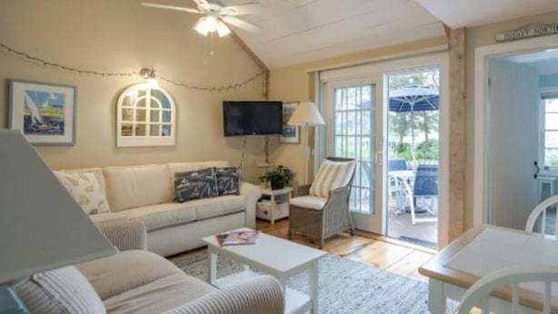 A cozy living room with a sofa, armchairs, a coffee table, and a TV. There is a ceiling fan, a window with curtains, and a door leading outside. - Cape Cod Cottage steps from Private Beach (Mashpee)