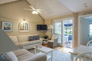 A cozy living room with a sofa, armchairs, a coffee table, and a TV. There is a ceiling fan, a window with curtains, and a door leading outside. - Cape Cod Cottage steps from Private Beach (Mashpee)