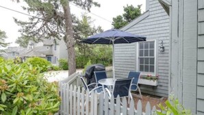 A patio area with a table and chairs, a barbecue grill, and a white picket fence. - Cape Cod Cottage steps from Private Beach (Mashpee)