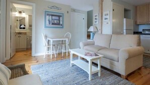 A living room with a beige sofa, a white coffee table, and a dining area with a white table and chairs. There is a kitchen area with cabinets and a refrigerator. - Cape Cod Cottage steps from Private Beach (Mashpee)