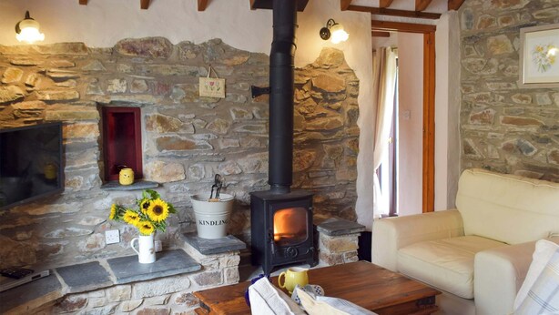 A cozy living room with a stone wall, a fireplace, a sofa, and a coffee table. - Old Chapel Cottage - Dinas Country Club (Dinas Cross, near Newport)