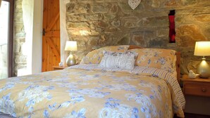 A bedroom with a stone wall, wooden beams, a bed with floral bedding, two bedside lamps, and a heart-shaped decoration. - Old Chapel Cottage - Dinas Country Club (Dinas Cross, near Newport)
