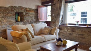 A cozy living room with a stone wall, a wooden coffee table, a beige sofa, and a window with curtains. - Old Chapel Cottage - Dinas Country Club (Dinas Cross, near Newport)
