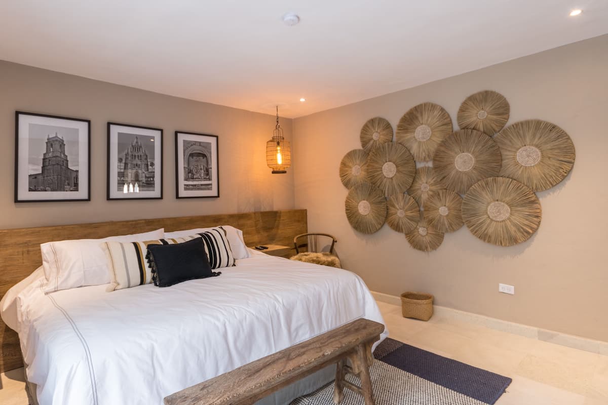 Une chambre à coucher avec un grand lit, une tête de lit en bois, un banc et des éléments décoratifs muraux, notamment des tableaux encadrés et des décorations murales tissées.