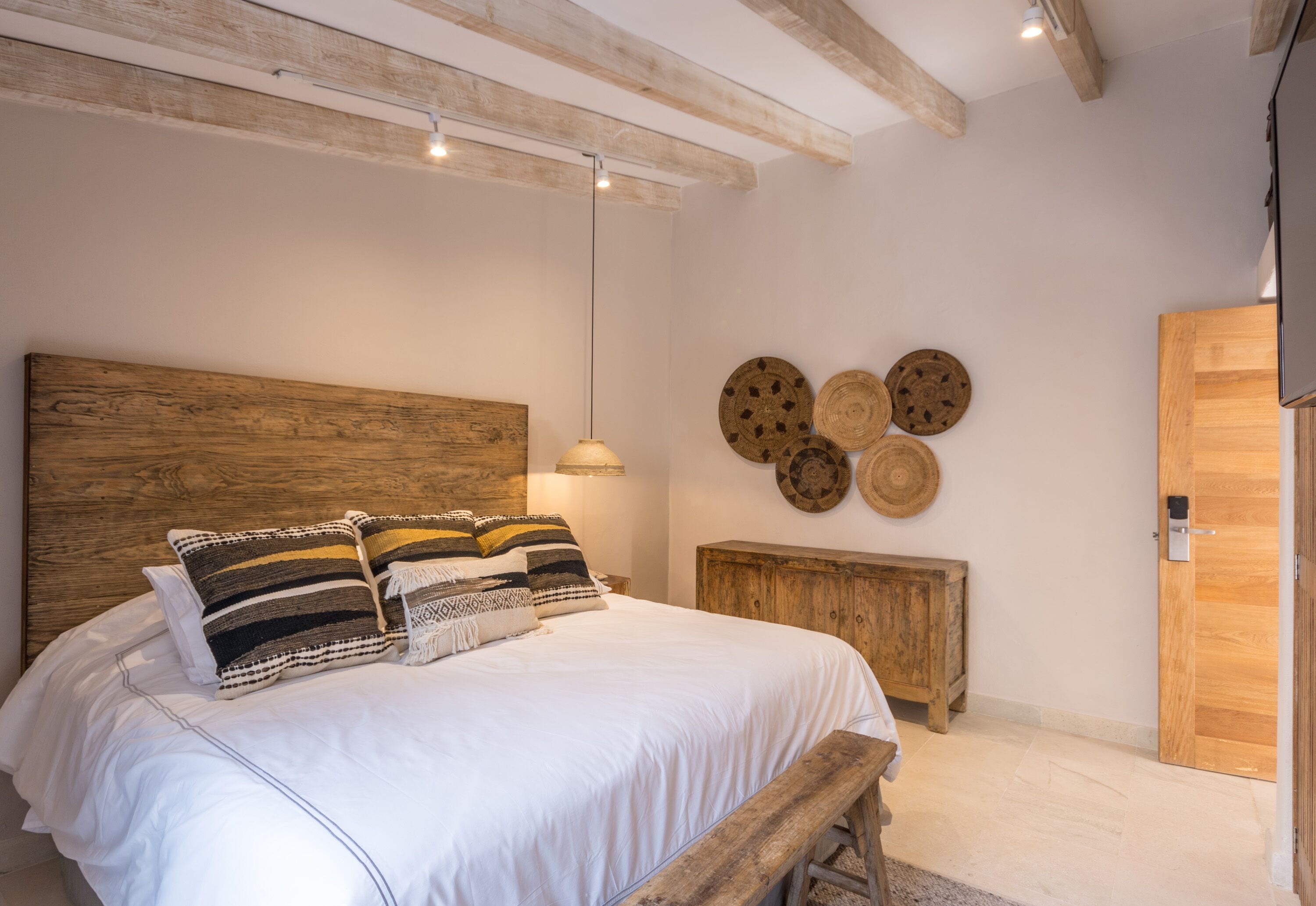 A bedroom with a wooden headboard, a white bedspread, and a wooden bench. There are wooden wall decorations and a wooden door.