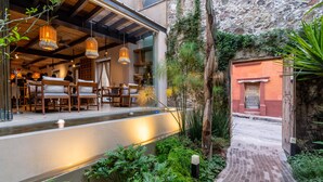 Un espace extérieur aménagé pour les repas, avec des tables et des chaises en bois, une fontaine ou un autre élément aquatique, et une végétation luxuriante.