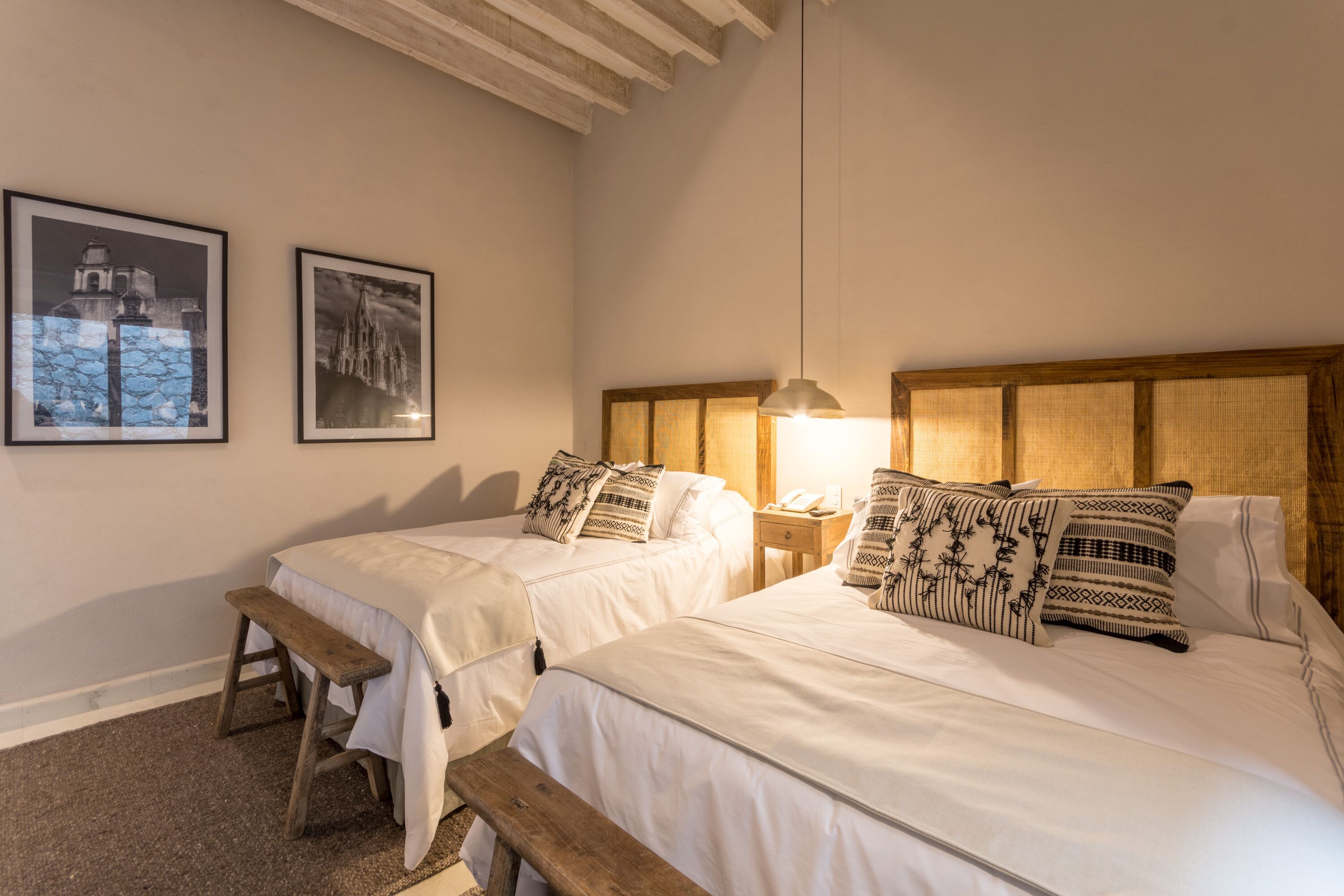 Une chambre avec deux lits, un banc en bois, une table de chevet avec une lampe et deux tableaux encadrés au mur.
