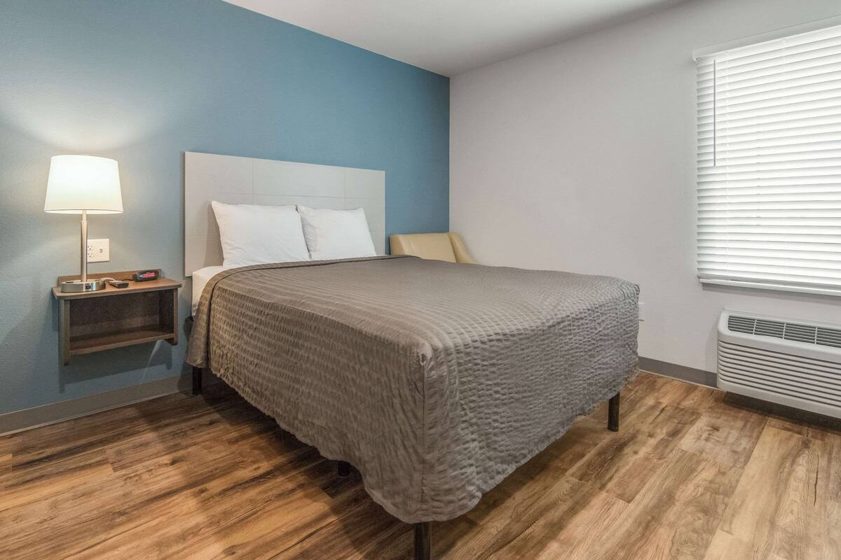 Une chambre d’hôtel avec un lit, une table de chevet, une lampe et une fenêtre avec des stores.
