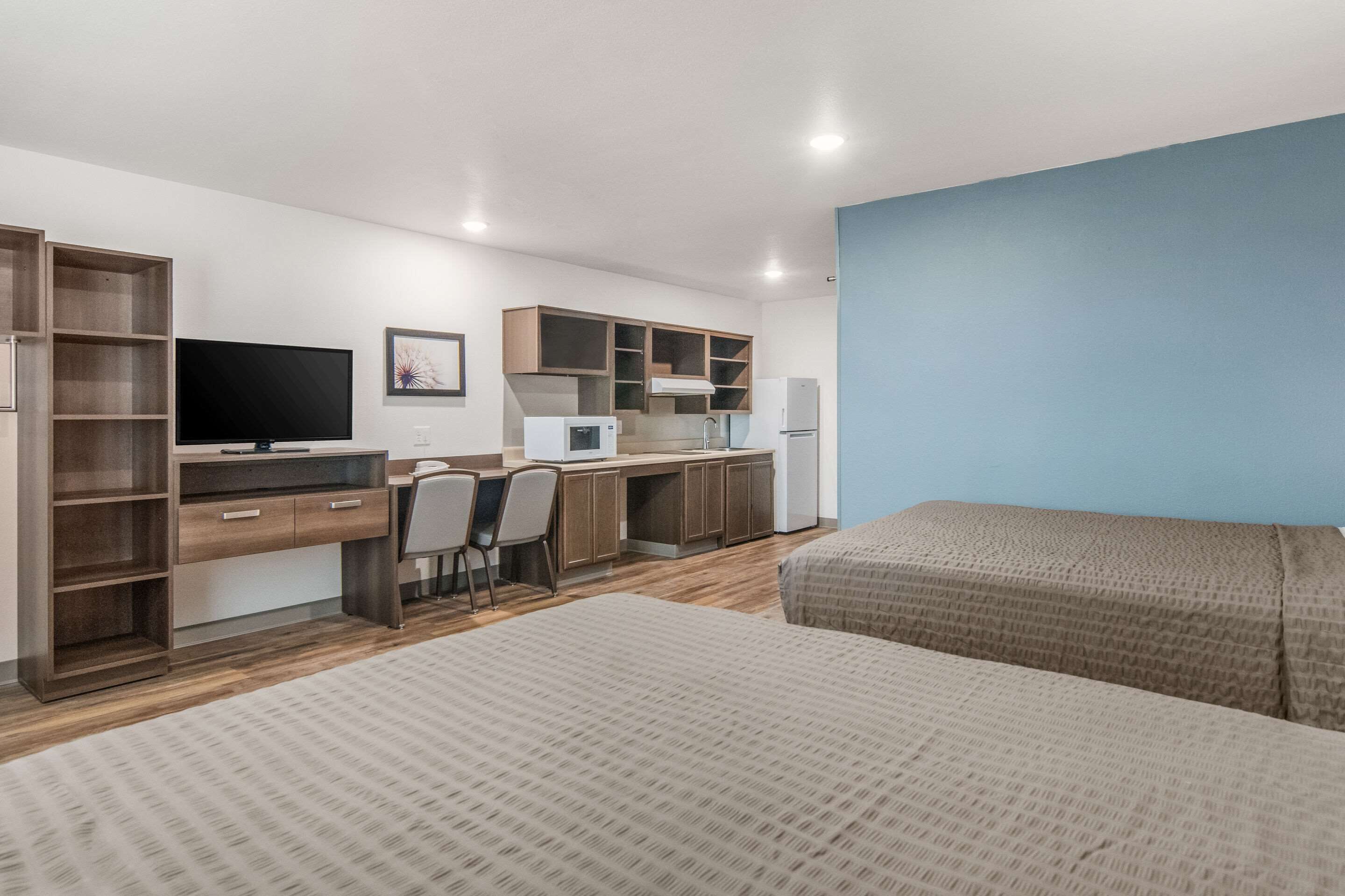 A modern hotel room with a large bed, a desk with a computer, a TV, and a kitchenette.