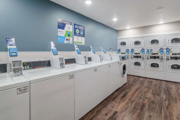 Un cuarto de lavandería con varias lavadoras y secadoras, estaciones de pago digital y carteles informativos en la pared.
