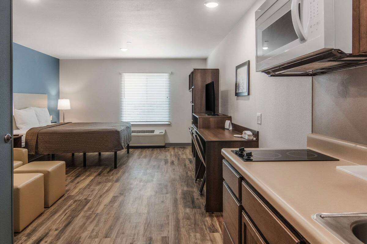 Une chambre d’hôtel compacte comprenant un lit, un petit coin salon, une kitchenette et une fenêtre avec des stores.