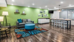 A modern living room with a green accent wall, a sofa, a blue ottoman, a dining area with a table and chairs, and a flat-screen TV mounted on the wall. - WoodSpring Suites North Ft Worth Alliance TX Speedway (Fort Worth)