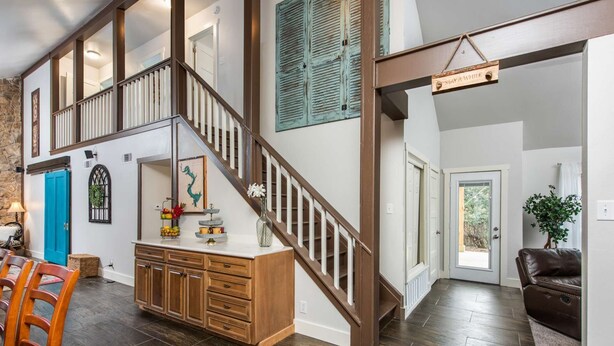 A spacious living area with a wooden staircase, a dining table with chairs, a kitchen area, and a view of the outdoors.