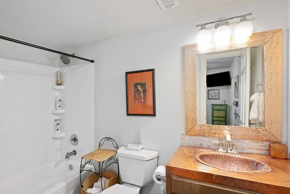 A bathroom with a white toilet, a wooden vanity with a copper sink, a large mirror with a textured frame, and a wall-mounted shelf with toiletries.