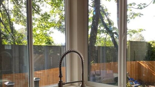 A kitchen with a double sink, a kettle, and a window overlooking a backyard with a wooden fence and trees. - Upscale Modern Private Cottage (Kirkland)