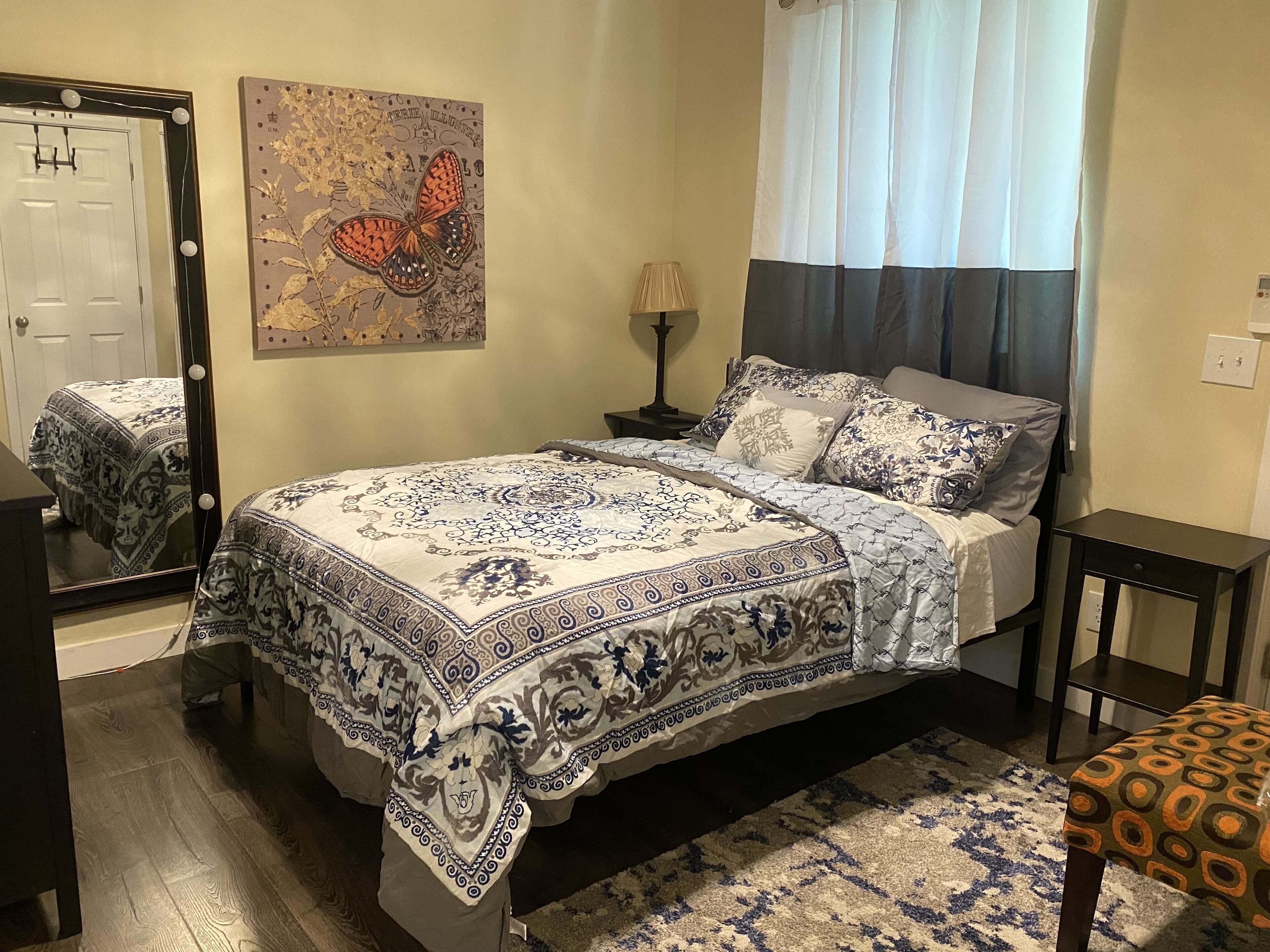 A bedroom with a bed, a nightstand, a mirror, a lamp, and a window with curtains.
