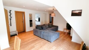 Un salon moderne avec un canapé d’angle gris, un plancher en bois, une porte en bois, une cuisine en arrière-plan et un luminaire suspendu.