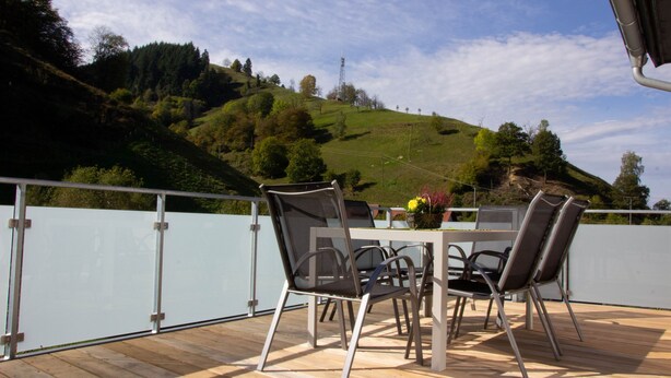 Une terrasse avec une table et des chaises, donnant sur un paysage vallonné parsemé d’arbres et sous un ciel dégagé.