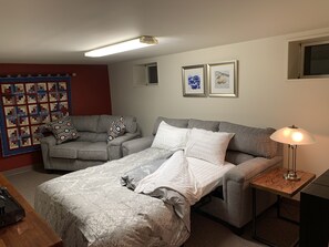 Quarto com cama, dois sofás cinzentos, mesa de cabeceira de madeira com abajur, painel de parede acolchoado e duas fotografias emolduradas na parede.