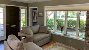 Ein Wohnzimmer mit einem beigen Sofa, einem kleinen Tisch und einem großen Fenster mit Blick auf Grünfläche.