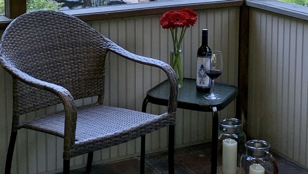 Ein Korbstuhl und ein kleiner Tisch mit einer Flasche und einem Glas, eine Vase mit einer roten Blume und Kerzen auf einer Veranda mit Blick auf einen Garten und einen Steinfall.