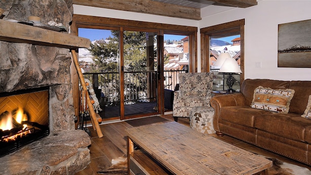 Una sala de estar acogedora con chimenea de piedra, una mesa de centro de madera, un sofá de cuero marrón y vistas a una montaña nevada a través de amplios ventanales.