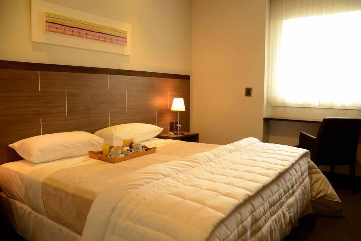 Ein ordentlich bezogenes Bett mit einem Frühstücksbrettchen, einer Nachttischlampe und einem Sessel in einem Hotelzimmer.