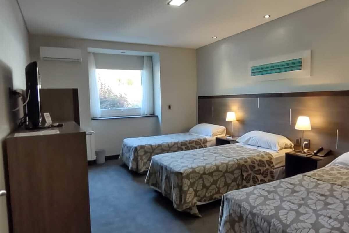 Ein Hotelzimmer mit drei Einzelbetten, einem Schreibtisch, einem Fernseher und einem Fenster mit Vorhängen.