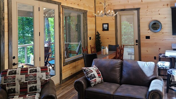 Een gezellig interieur van een chalet, met een houten plafond, een plafondventilator, een kroonluchter, een leren bank, een bijzettafeltje en een eethoek met stoelen en een tafel.