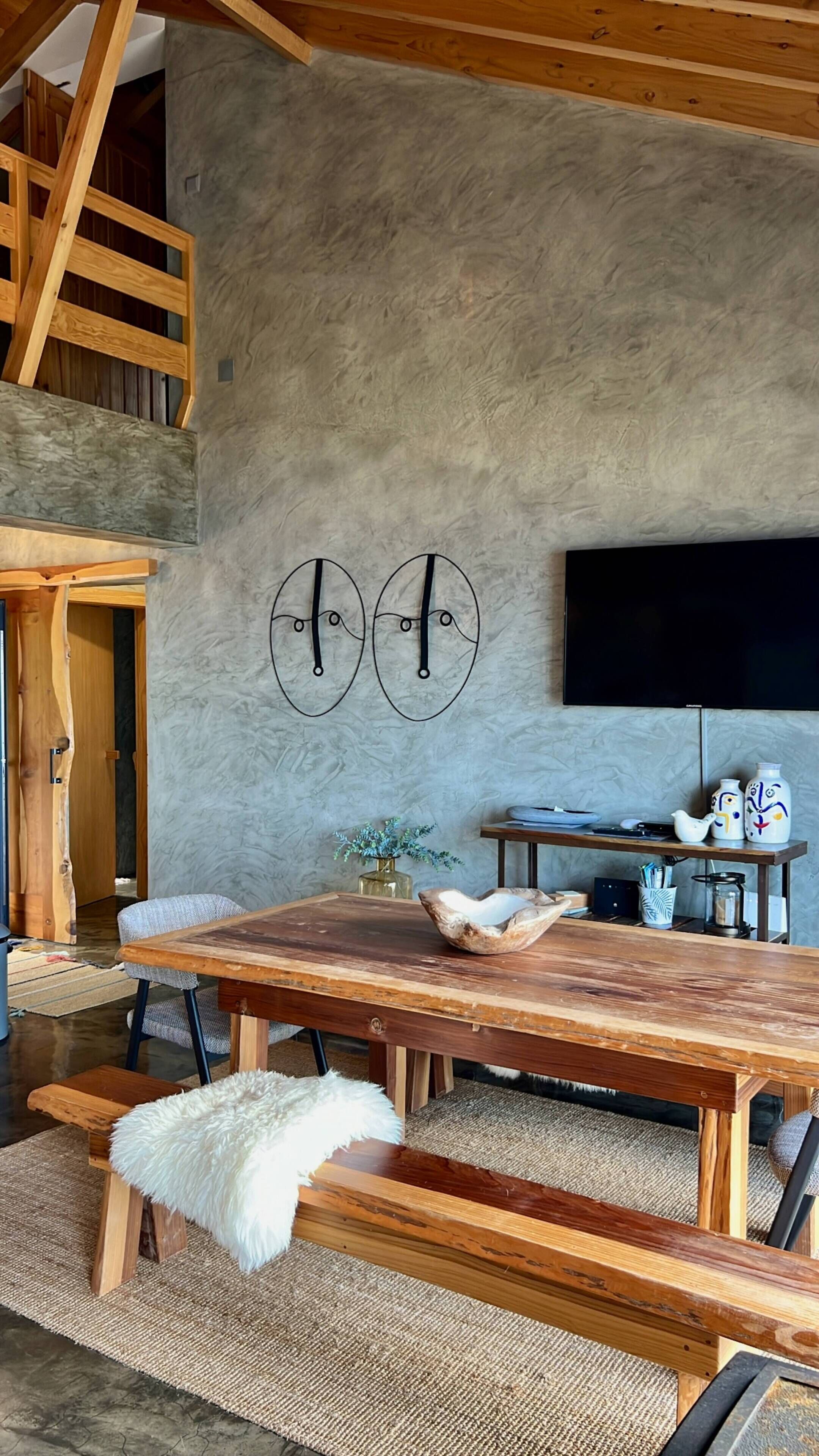 A rustic interior with a wooden dining table, benches, a flat-screen TV mounted on a textured wall, and a staircase leading to an upper level.