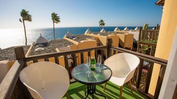 Een balkon met een glazen tafel en twee witte stoelen, met uitzicht op een strand met palmbomen en een heldere lucht.