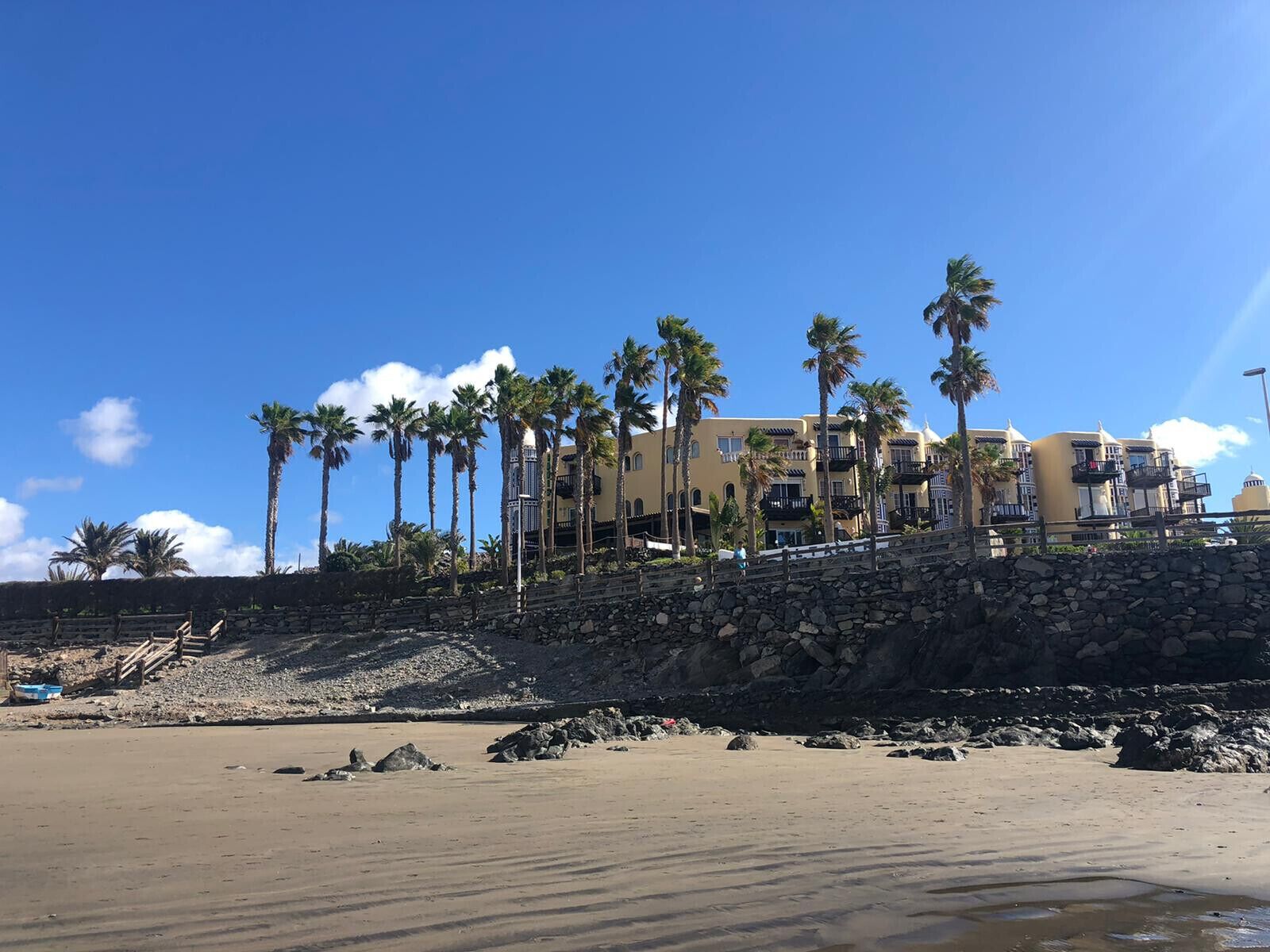 Hotel aan het strand met een stenen steunmuur, palmbomen en een helderblauwe lucht.